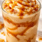 Tall glass of thick caramel ribbon crunch frappuccino topped with whipped cream, caramel drizzle, and golden crunch pieces.